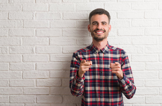 Young adult man standing over white brick wall pointing fingers to camera with happy and funny face. Good energy and vibes.
