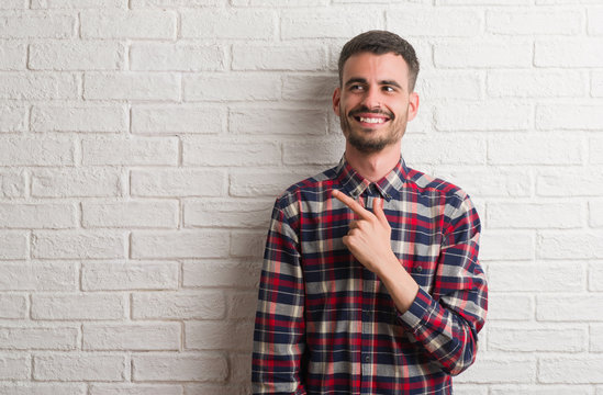 Young adult man standing over white brick wall cheerful with a smile of face pointing with hand and finger up to the side with happy and natural expression on face looking at the camera.