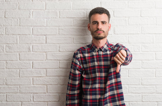 Young adult man standing over white brick wall looking unhappy and angry showing rejection and negative with thumbs down gesture. Bad expression.