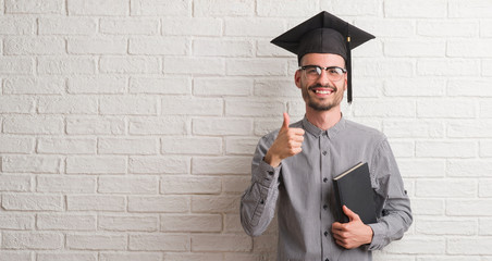 Young adult man over brick wall wearing graduation cap happy with big smile doing ok sign, thumb up...