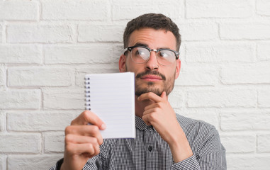 Young adult man holding notebook standing over white brick wall serious face thinking about...