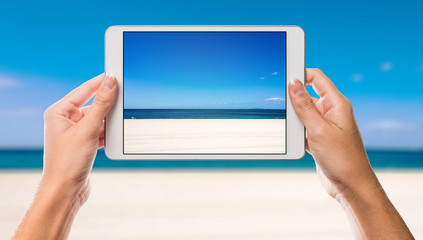 Hands holding tablet with holiday beach and sea on screen and beach and coast background