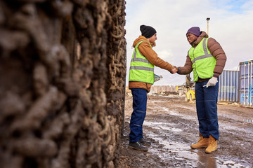 Full length portrait of two industrial workers wearing reflective jackets shaking hands outdoors standing by big dirty truck