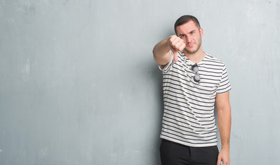 Young caucasian man over grey grunge wall looking unhappy and angry showing rejection and negative with thumbs down gesture. Bad expression.