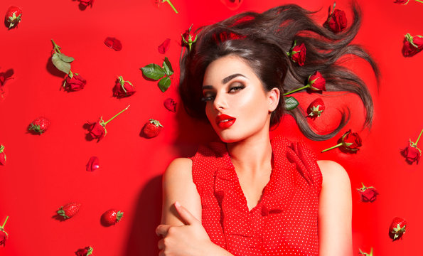 Beauty sexy model girl lying on red background with rose flowers and strawberries. Beautiful brunette young woman with long hair and perfect make-up, red seductive lips, smoky eyes. Trendy makeup