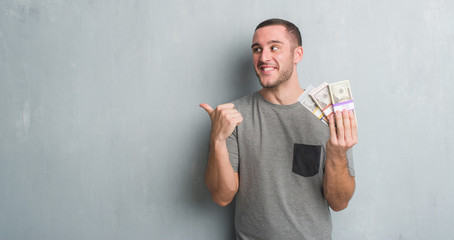 Young caucasian man over grey grunge wall holding a bunch of money pointing and showing with thumb...