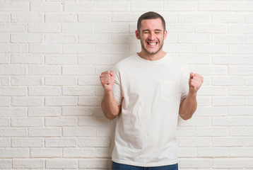 Young caucasian man standing over white brick wall excited for success with arms raised celebrating victory smiling. Winner concept.