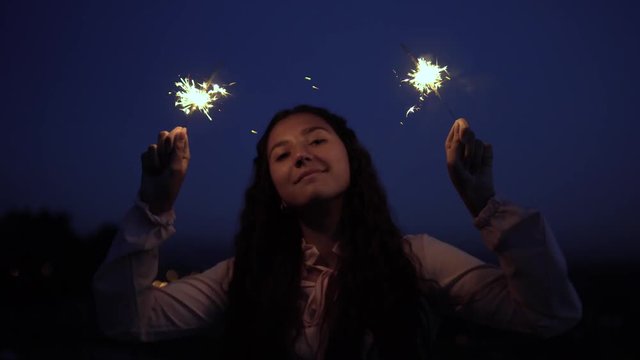 A young girl with long dark hair stands against the background of the city at night and holds fireworks in a good mood. slow motion. 4K