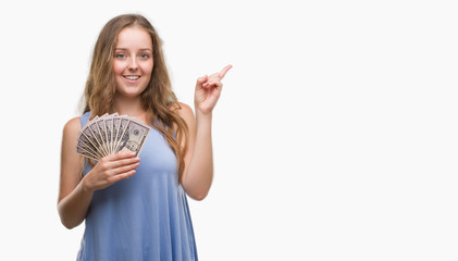 Young blonde woman holding dollars very happy pointing with hand and finger to the side