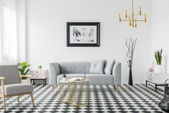 Real photo of a modern living room interior with a grey sofa, armchair, painting, golden lamp and tiles on the floor