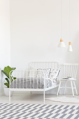 White chair next to bed in bedroom interior with patterned carpet and plant. Real photo