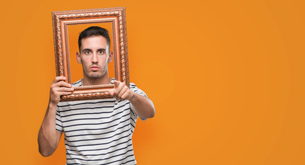 Handsome young man looking through vintage art frame pointing with finger to the camera and to you, hand sign, positive and confident gesture from the front