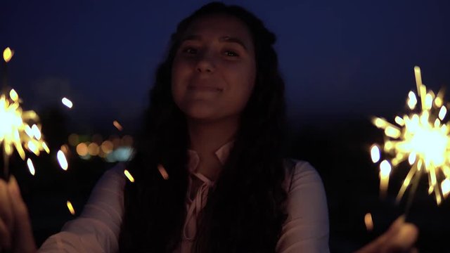 A young woman with long dark hair holds fireworks at night in the background of the city and is happy to have a good mood. slow motion. Portrait. 4K