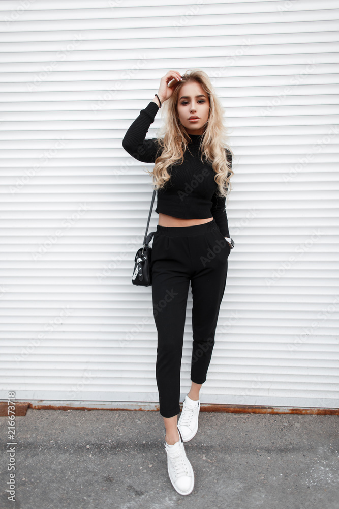 Wall mural Young beautiful fashion model girl with a handbag in a black T-shirt, pants and white sneakers poses near a white metal wall