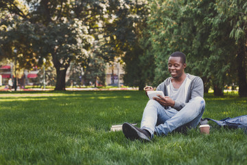 Naklejka na ściany i meble Happy african-american studen sitting with notebook on grass