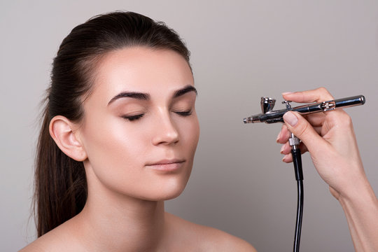 Makeup Artist Using Airbrush. Fashion Brunette Model With Hair Tail Over Gray Background. Professional Makeup With Aerograph. Girl Doing Makeup In Beauty Salon. Female Face With Natural Clean Skin.