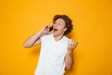 Photo of happy young guy with curly hair shouting and talking on smartphone, isolated over yellow background