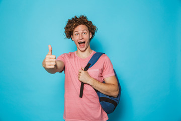 Photo of student guy with curly hair wearing casual clothing and backpack smiling and showing thumb up, isolated over blue background