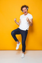 Full length photo of young smiling guy with curly hair listening to music via headphones and singing, isolated over yellow background