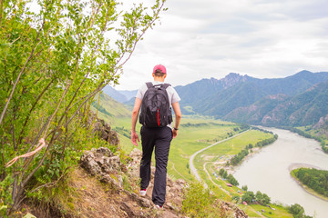 Naklejka premium A young man in a cap with a backpack and black sports pants is going on the edge of a cliff in the mountains of the Altai, from where he opens a view of the river Katun and the island