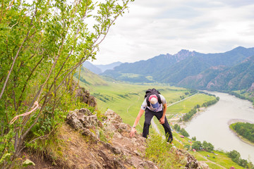 A young guy in a cap with a backpack and black sports pants is climbing up on the edge of a cliff in the mountains of the Altai, from where he opens a view of the river Katun and the island