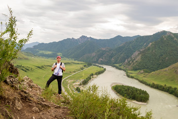 A young guy in a cap with a backpack and black sports pants is standing on the edge of a cliff in the mountains of the Altai, from where he opens a view of the river Katun and the island