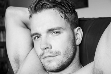 Hunky muscular, sexy shirtless man with muscules and pecs sitting on sofa, looking at camera