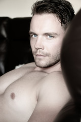 Hunky muscular, sexy shirtless man with muscules and pecs sitting on sofa, looking at camera