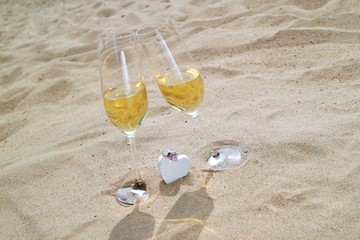 Zwei Gl&auml;ser mit Sekt stehen im Sand ein Dekoherz mit Ring liegt dazwischen 