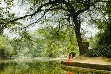 Under large tree a girl sits and studies yoga in the lotus poses. Hands raised up. Near the lake with a beautiful reflection of the tree. Gardens of Lodi, India. Advertising of yoga tours. Meditate.