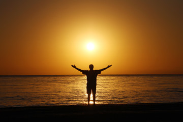 A man on the beach welcomes the sunrise. Hands are spread apart.