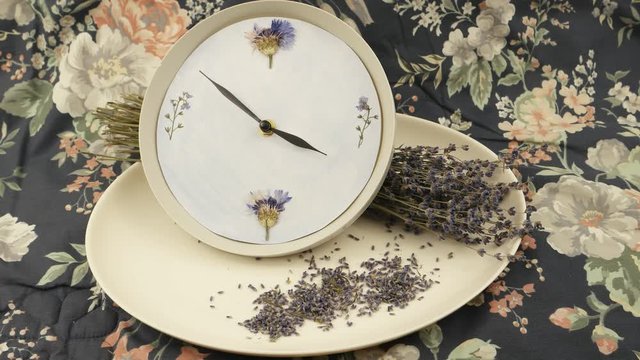 A clock, a white faience dish, and bouquet of dried lavender against the background of fabric with a floral pattern, floristic composition
