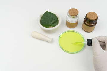 herbal medicine research concept. the scientist holding a green liquid dropper on watch glass, an organic green leaf in mortar, a pestle and bottles on the white table in laboratory