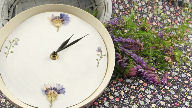 A clock, a blue wicker basket and a small bouquet of wild flowers against the background of fabric with a floral pattern, floristic composition