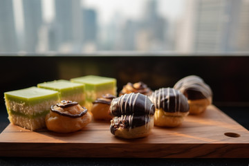 Assortment of desserts on wooden tray. Tasty and delicious layer cake and Cream Puff or profiterole with chocolate decoration against cityscape urban skyline background at rooftop bar.Selective focus