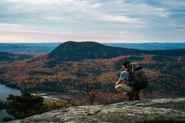 A man with a backpack kneels on top of a mountain in Maine in the fall with autumn foliage and a...