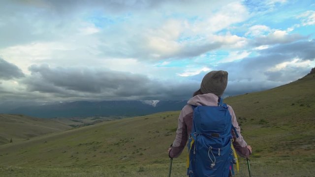 A girl with a backpack and trekking sticks stands and admires the beautiful mountain view that has opened. The valley, with low clouds covering the snow-capped mountains. Hiker trekking or walking of