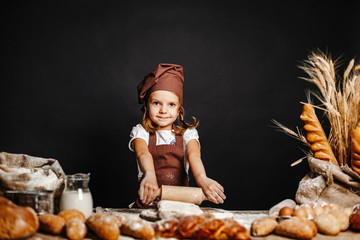 Charming little girl in apron and hat standing at table kneading bread dough and having fun spending time cooking