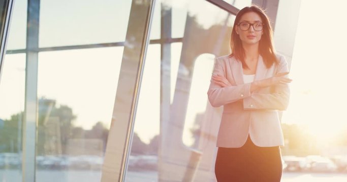 Portrait of the attractive Caucasian woman standing outside big office building and turning her head to the camera, crossing hands and smiling in the sunlight.