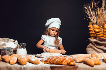 Charming little girl with curly hair in white apron and hat standing at table kneading bread dough...