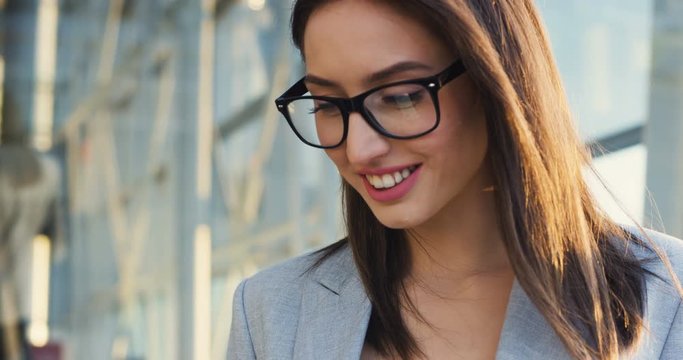 Close up of the Caucasian young woman in business style taping and texting on the smartphone device, then smiling nice as reading something pleasent.