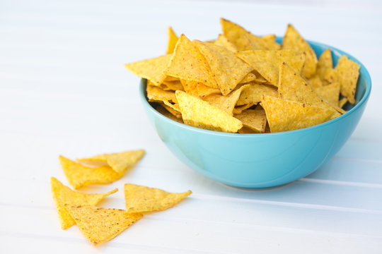 Yellow triangle shaped crispy nachos in a blue turquoise bowl placed on white table, copy space, some nachos laying out