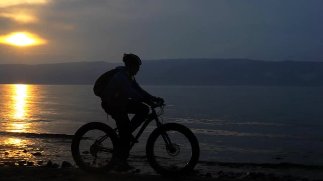 Fat bike also called fatbike or fat-tire bike in summer riding in the beach. The athlete passes in a frame silhouette against a beautiful sunset on the sea. Rides directly against the background of