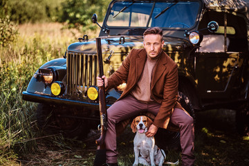 Hunter in elegant clothes holds a shotgun and sits together with his beagle dog while leaning...