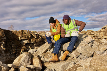 Full length portrait of two industrial  workers wearing reflective jackets, one of them African, relaxing taking coffee break from work  and chatting on mining worksite outdoors