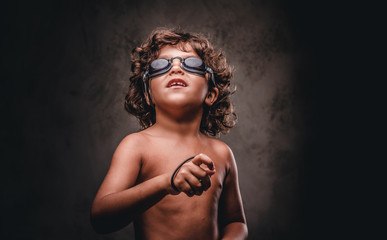 Funny little shirtless boy in swimming goggles in a studio. Isolated on the dark textured background.