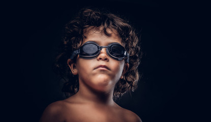 Cute little shirtless boy in swimming goggles posing in a studio. Isolated on the dark textured background.