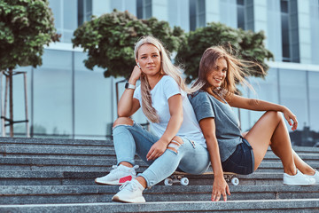 Portrait of a two young hipster girls sitting together on skateboard at steps on a background of the skyscraper.