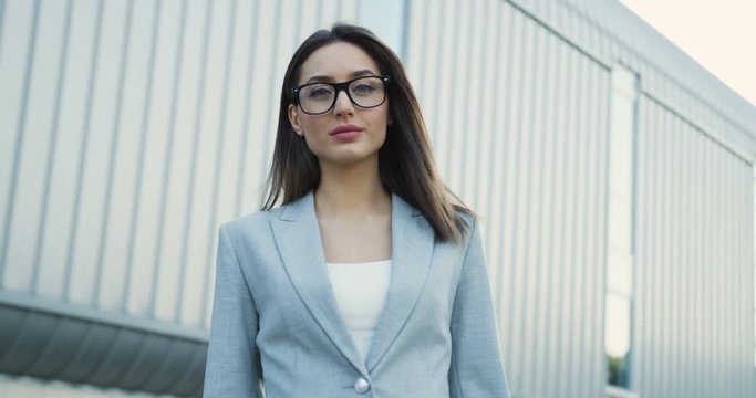 Portrait of the beautiful young businesswoman in the glasses turning her head to the camera near the big office building and smiling.