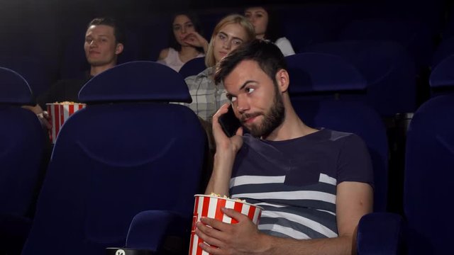 Young woman annoyed with a man talking on his phone during the movie at the cinema. Male spectator answering his phone at the movie theatre. Technology, multitasking, annoying concept.